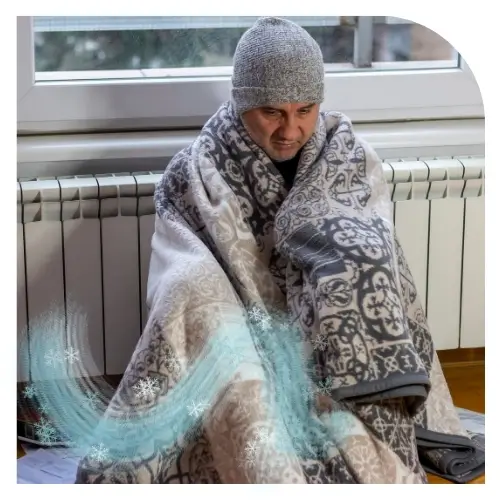 Ein Mann, der sich mit einer Decke wärmt und neben der Heizung sitzt, weil er keine abgedichteten Fenster hat.